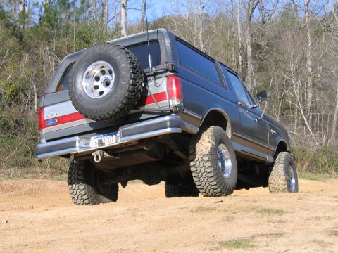 1989 Ford Bronco - This lifted Ford Bronco is shown from the rear, parked on a dirt path with trees in the background.