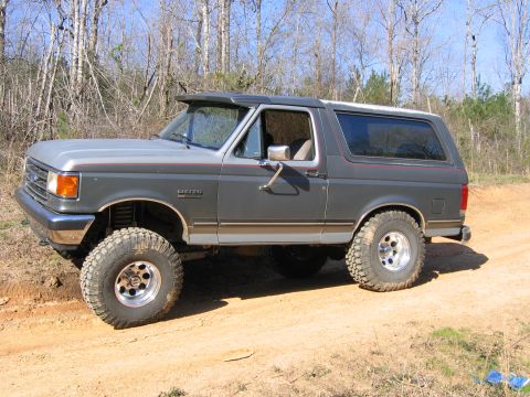 1989 Ford Bronco - This lifted Ford Bronco is parked on a dirt road with trees in the background.
