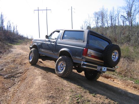 1989 Ford Bronco - This lifted Ford Bronco is navigating a rough, unpaved trail with trees and power lines in the background.