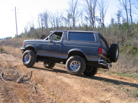 1989 Ford Bronco - A dark gray Ford Bronco navigates an off-road trail with large tires and a rear-mounted spare.