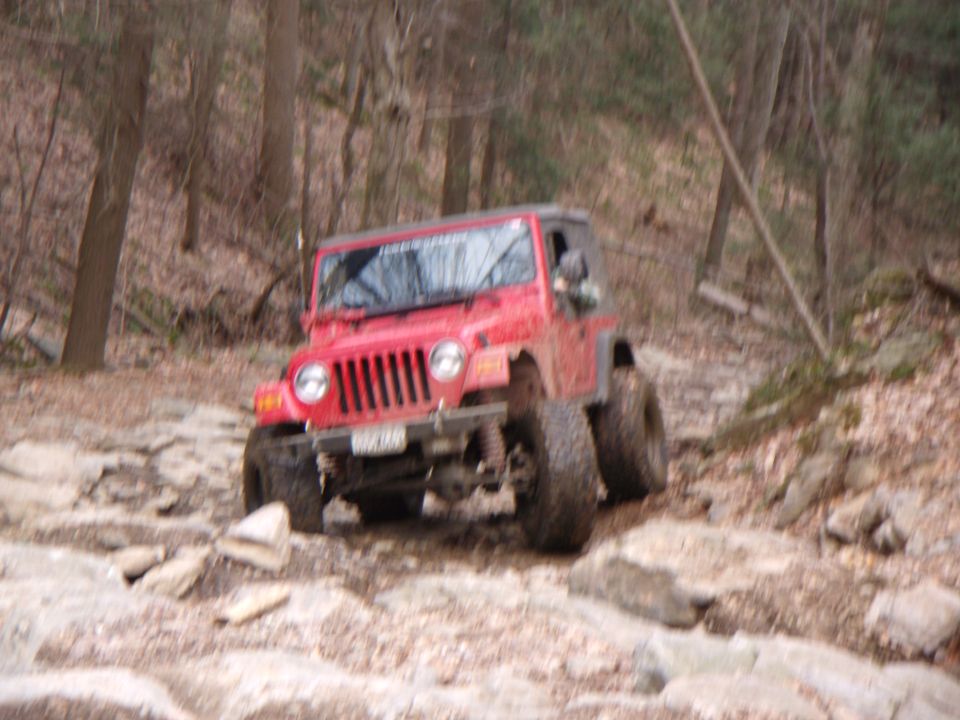 Red 2003 Jeep Wrangler navigating a rocky trail with trees in the background