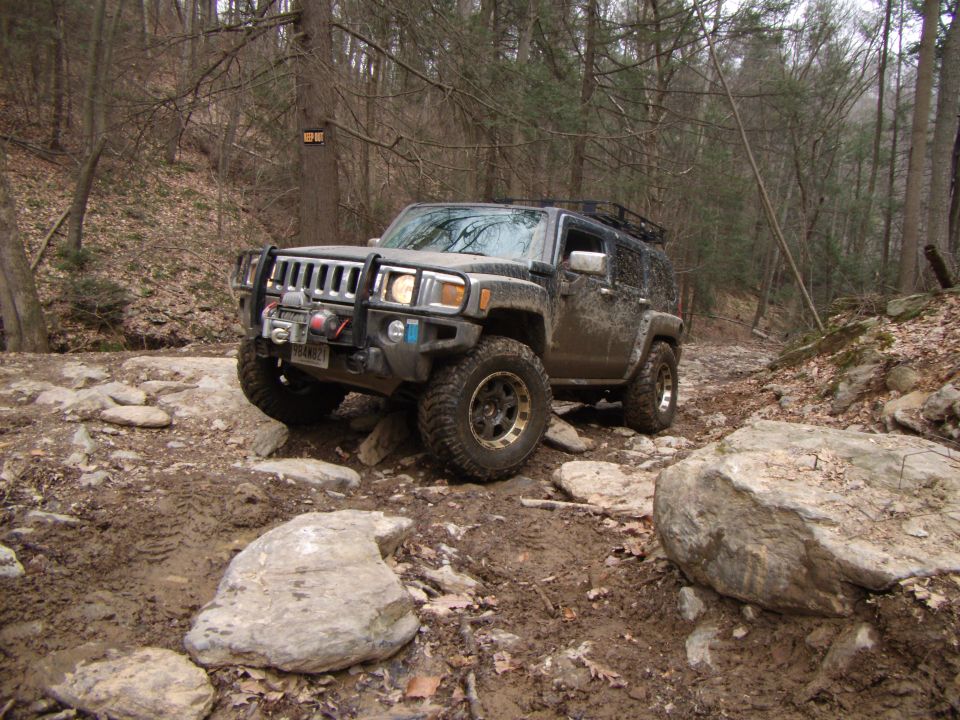 Hummer H3 driving over rocks and mud on a wooded trail. It has a winch and roof rack