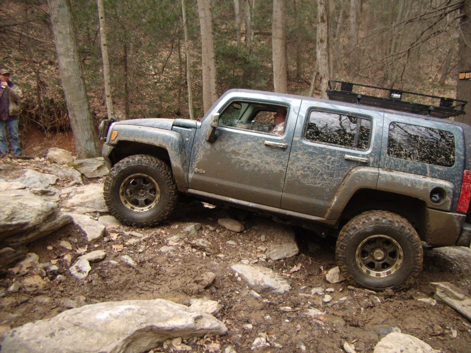 Mud-splattered dark gray Hummer H3 off-roading on a rocky, wooded trail