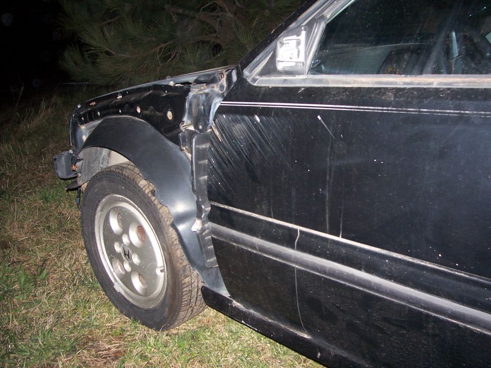 Black coupe at night with front fender removed, alloy wheel and door visible on grass