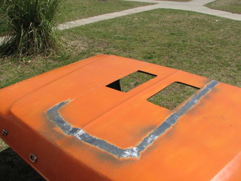 1979 Ford Bronco - Orange car hood with cutouts and welded repair work, viewed outdoors.