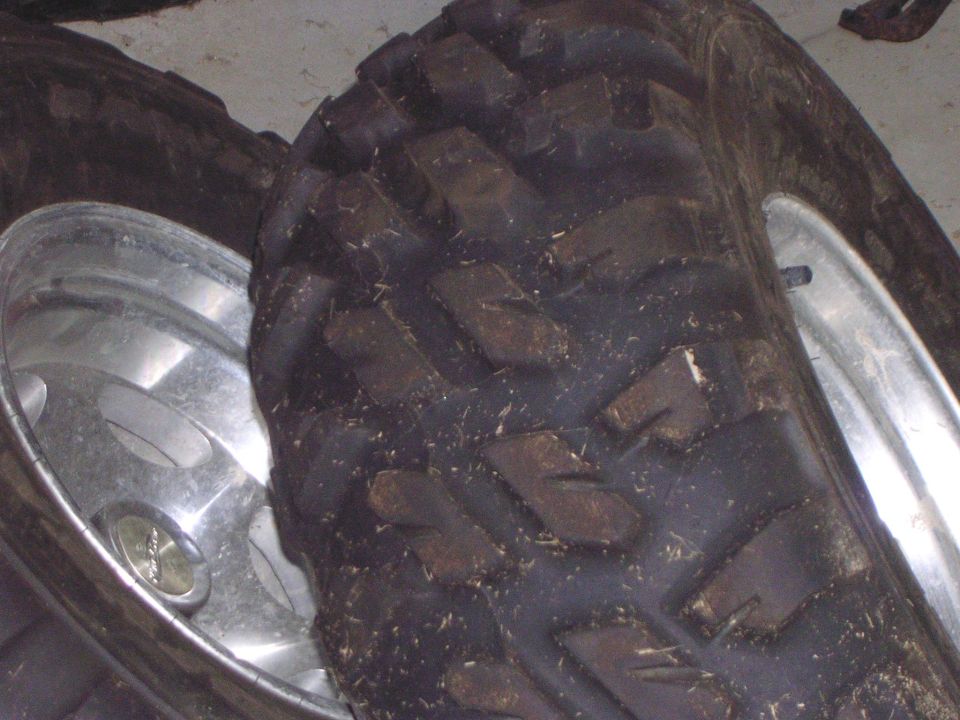 Two muddy knobby ATV tires on polished aluminum wheels stacked on a concrete floor