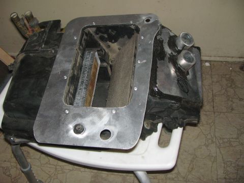 1990 Ford Bronco - disassembled black HVAC heater core box fitted with an aluminum mounting plate, resting on a white plastic chair.