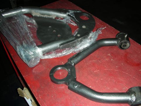 1998 Ford Crown Victoria - Tubular black-coated control arms and mounting brackets, partly wrapped in plastic, on a red workbench.