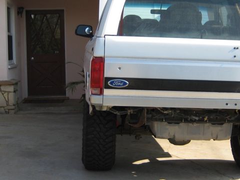 1996 Ford Bronco - Rear three-quarter view of a white Ford Bronco with a dark two-tone lower body stripe and Ford oval badge, lifted on large mud-terrain tire…
