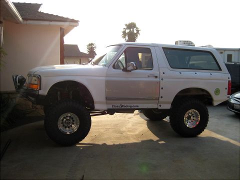 1996 Ford Bronco - A white two-door Ford Bronco SUV shown in side profile with a heavy lift kit, oversized mud-terrain tires and polished beadlock-style wheel…