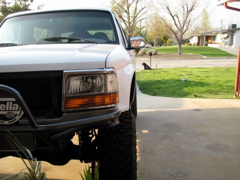 1996 Ford Bronco - Close front view of the driver-side corner of a white lifted Ford Bronco parked on a driveway, showing a clear headlight with amber turn si…