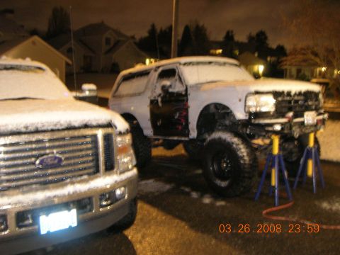 1996 Ford Bronco - A white Ford Bronco sits on jack stands in the snow, its driver's door removed.