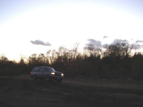 1989 Ford Bronco - A Ford Bronco is seen driving on a dirt path during sunset with trees lining the horizon.