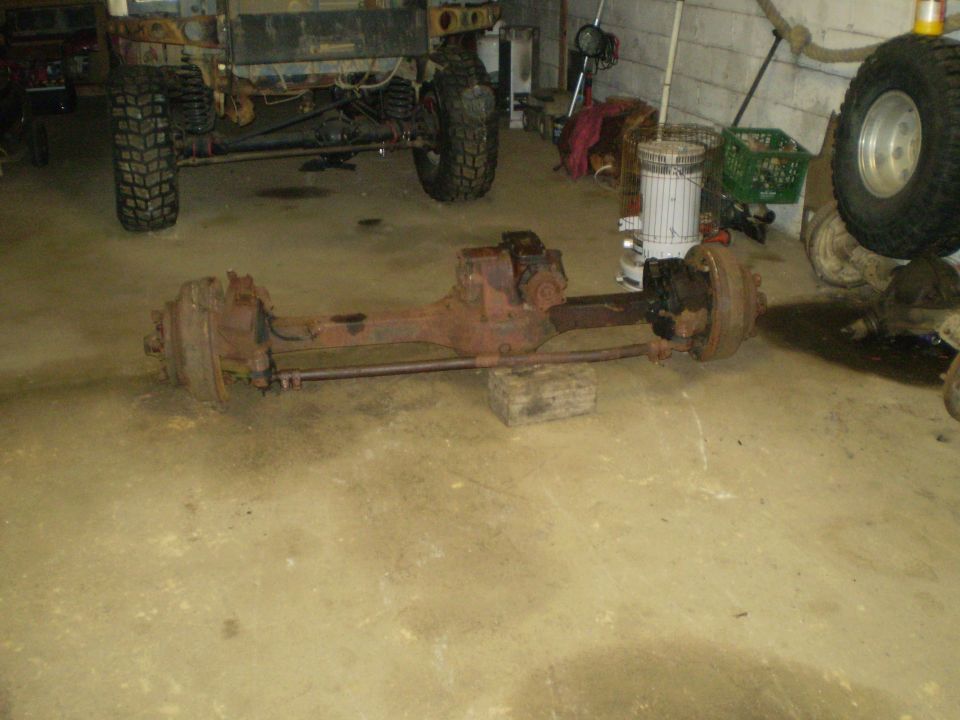 Front axle assembly removed from a vehicle, sitting on a concrete floor. Another vehicle is partially visible in the background
