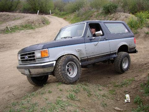 1989 Ford Bronco - A two-tone blue and silver Ford Bronco navigates a dirt path, with a driver in the cabin.