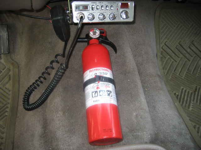 Red Kidde fire extinguisher on vehicle carpet beside a Uniden CB radio and coiled mic