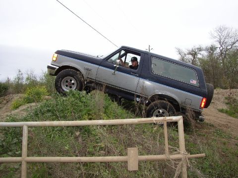 1989 Ford Bronco - A two-tone blue and silver Ford Bronco navigates an off-road trail, viewed from the front-right side.