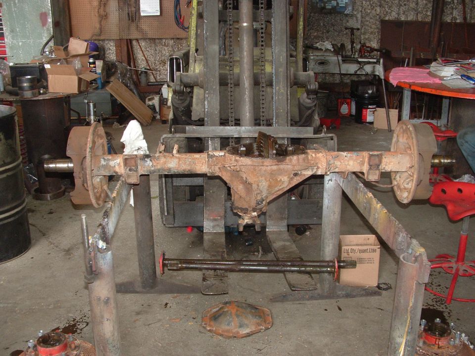Rusty rear axle assembly with differential cover removed, on a stand in a workshop in front of a forklift