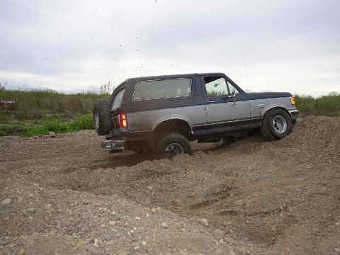 1989 Ford Bronco - A two-tone blue and silver Ford Bronco navigates a muddy terrain, with its rear tire partially submerged.