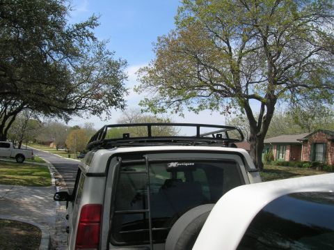 1999 Land Rover Discovery II - Rear view of a silver SUV equipped with a roof rack and ladder, parked next to another white vehicle.