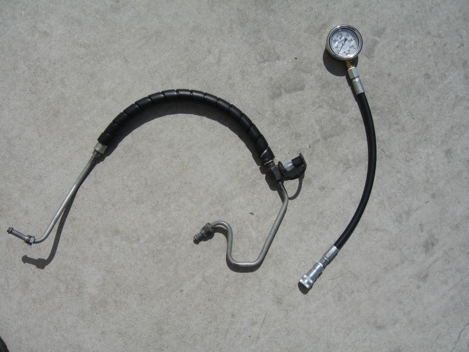 Power steering hose and pressure gauge on concrete. Hose has black protective wrap. Gauge reads approximately 500 psi