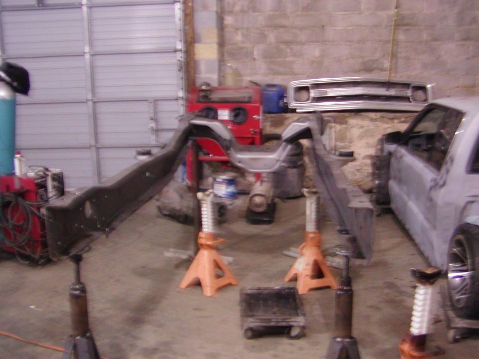 Vehicle frame on orange jack stands in a garage, with a partial vehicle body to the right