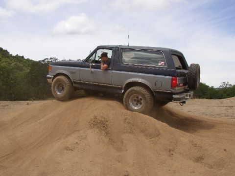 1989 Ford Bronco - A man drives a two-tone Ford Bronco up a sandy incline under a cloudy sky.