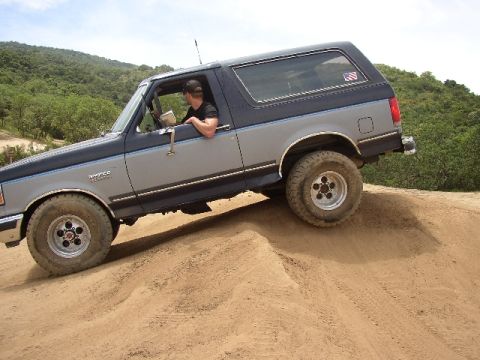 1989 Ford Bronco - A two-tone grey and blue SUV navigates a sandy incline, with the "Bronco" model name visible on its side.