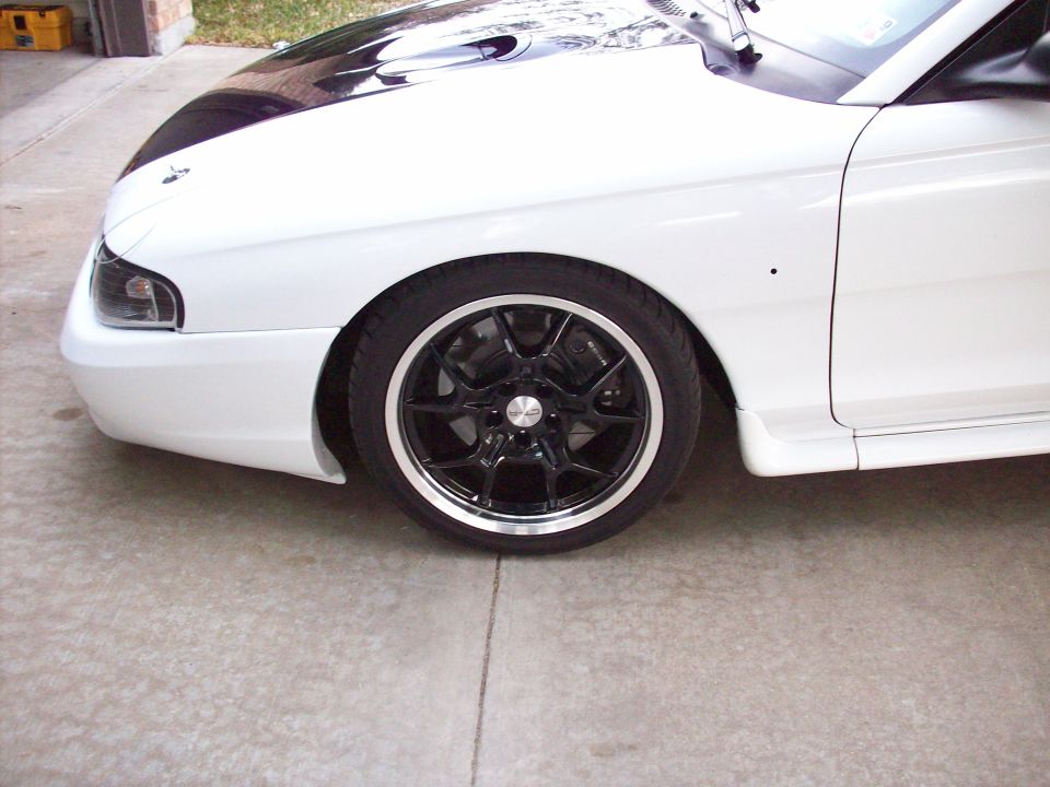 Front fender and wheel of a white 1997 Ford Mustang with black racing stripes on the hood