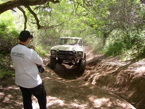 1989 Ford Bronco - Off-road vehicle navigates a challenging dirt trail surrounded by trees. A person stands in the foreground.
