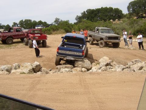 1989 Ford Bronco - Several trucks are off-roading on a dirt course with large rocks.