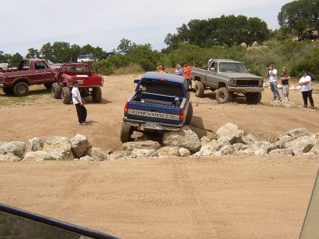 Blue Chevy pickup climbing rocks at an off-road park with lifted trucks and spectators