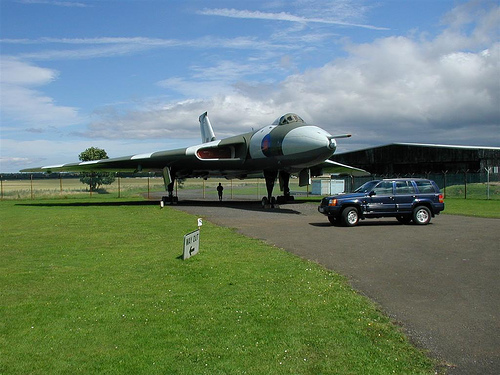 Blue SUV parked on asphalt next to a large jet airplane on a grassy field under a cloudy sky