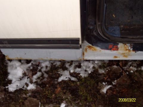 1988 Ford Bronco - Low close-up of the rocker panel and lower door jamb on the passenger side of a white-and-dark-blue truck, showing rust bleeding through th…