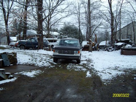 1988 Ford Bronco - A dark pickup truck is parked in a snowy yard with other vehicles and equipment.