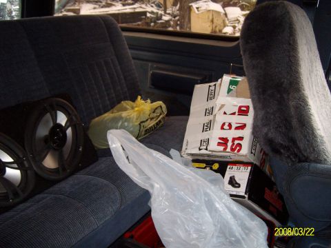 1988 Ford Bronco - Cluttered rear interior of a truck-based SUV with blue cloth bench seat and a gray sheepskin-covered front seat back at right. Loose afterm…