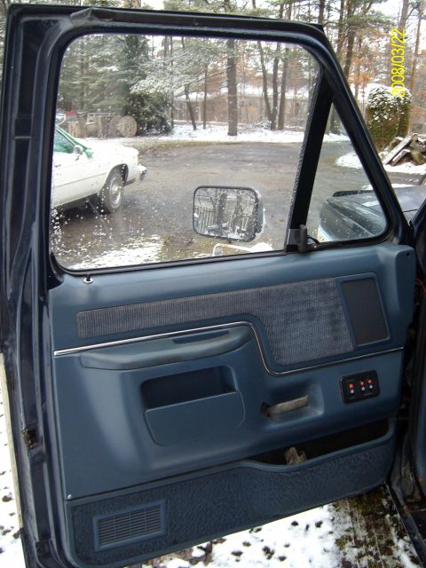 1988 Ford Bronco - Open driver's door of a dark blue truck photographed from outside, showing the blue interior door panel with cloth insert, chrome trim stri…