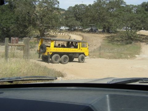 1989 Ford Bronco - A yellow, six-wheeled all-terrain vehicle drives on a dirt road, with other vehicles and trees visible in the distance.