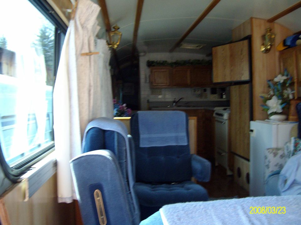 Interior of a converted bus camper with blue captain's chairs, oak cabinets and a range