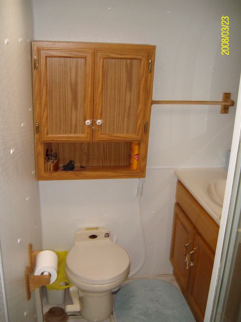 1988 Ford Bronco - Interior bathroom of an RV or travel trailer, showing a beige toilet with a wooden paper holder, an oak overhead cabinet with a latched doo…