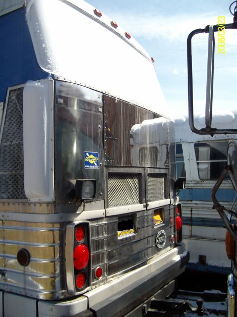 1988 Ford Bronco - Rear three-quarter view of a vintage stainless-and-blue coach bus in a snowy yard, showing the ribbed polished rear panel, round red tail l…