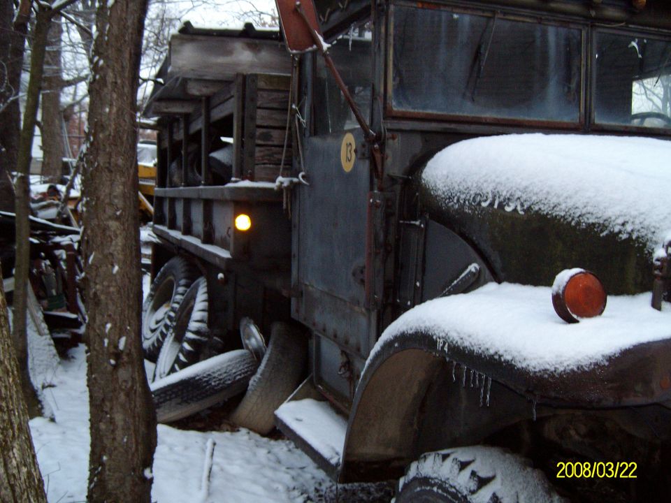 Old dump truck covered in snow, parked among trees. Visible details include wooden bed, large tires, and an orange marker light