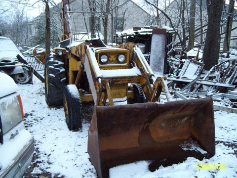 1988 Ford Bronco - Front view of a weathered yellow industrial tractor-loader backhoe parked in a snowy yard, its rusty front bucket lowered toward the camera…