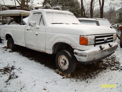 1988 Ford F-150 - A white pickup truck sits in the snow, its body showing signs of rust and being elevated by cinder blocks.