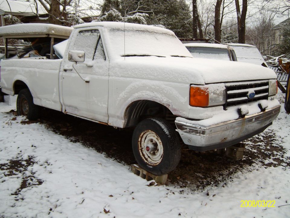 White pickup truck covered in snow, front quarter view. Rusty body, raised on cinder blocks