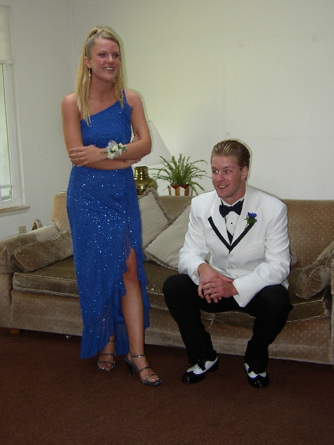 Blonde woman in blue gown standing beside a man in a white tuxedo on a couch