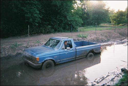 Blue single-cab pickup in a muddy puddle on a dirt lot