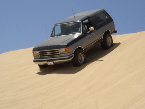 1989 Ford Bronco - A two-tone Ford Bronco navigates a sandy incline under a clear blue sky.