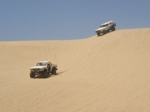 1989 Ford Bronco - Two trucks navigate sandy terrain on a bright, clear day.