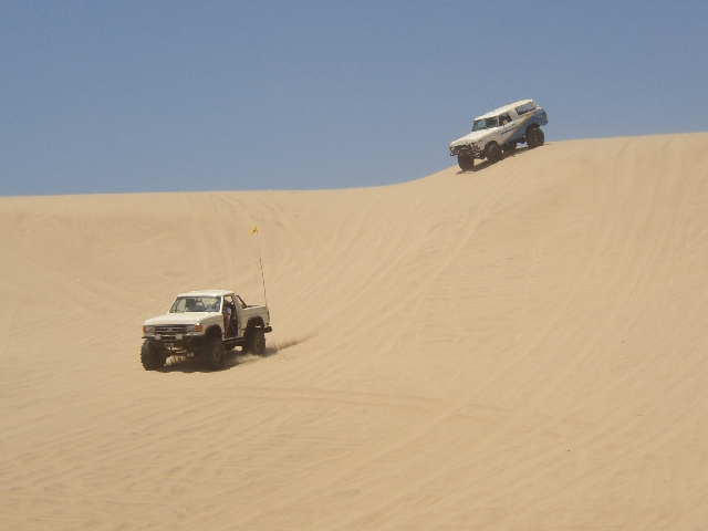 Two white lifted 4x4s on sand dunes, pickup with flag and SUV on ridge